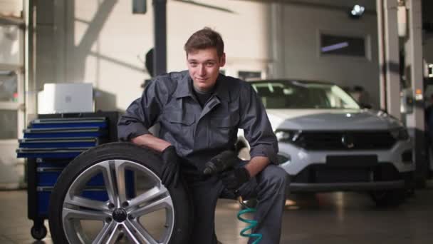 Portrait of a male tire specialist in a working uniform in a car service checks tires background of a car, smiles and looks at camera — Stockvideo