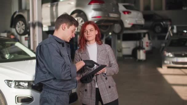Estação de serviço, um jovem profissional mostra um cliente feminino satisfeito em um tablet a condição técnica de um carro, sorri e olha para a câmera — Vídeo de Stock