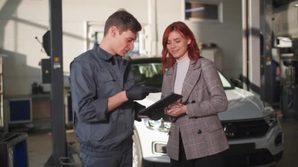 Service center, a young male mechanic shows a satisfied female client on a tablet technical condition of car during vehicle technical inspection — Vídeos de Stock