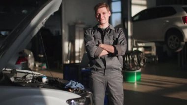 Üniformalı genç erkek uzmanın portresi açık kaputun yanında duruyor. Araba servis istasyonunda gülümsüyor ve kameraya bakıyor.