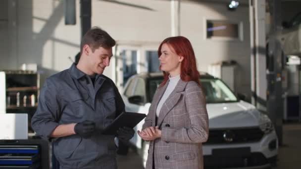 Automobile maintenance, male mechanic uses modern technology and shows technical moment of car on tablet beautiful female client, smiles and looks at camera — Vídeos de Stock