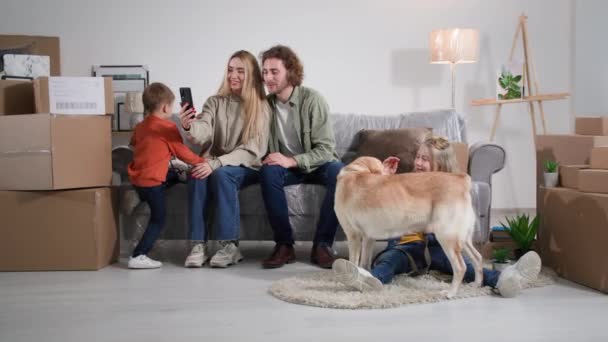 携帯電話を使ってネットで話しているポリエチレンで覆われたソファに座っている夫と妻子供とラブラドールの犬は家の温暖化で喜びます — ストック動画