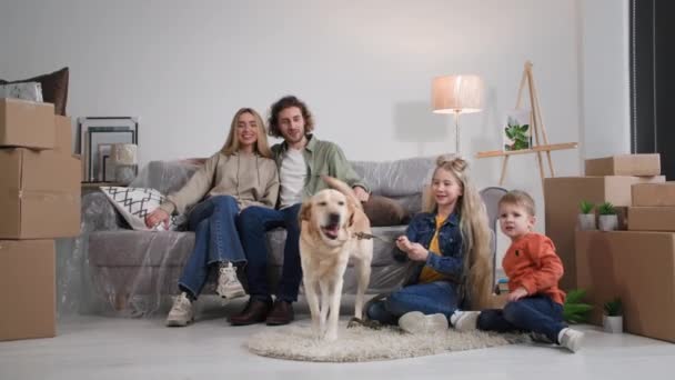 引っ越し後の家族の肖像画はアパートの購入を楽しんで子供たちは母親と父親の近くのラブラドール犬と遊ぶ新しい家のソファに座って — ストック動画