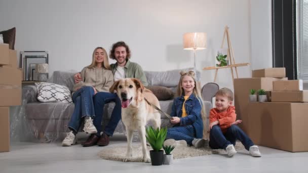 家族は新しい家に移りました息子娘とラブラドールの犬が段ボール箱に囲まれたソファに座っている間に抱きついた若いカップルの肖像画です — ストック動画