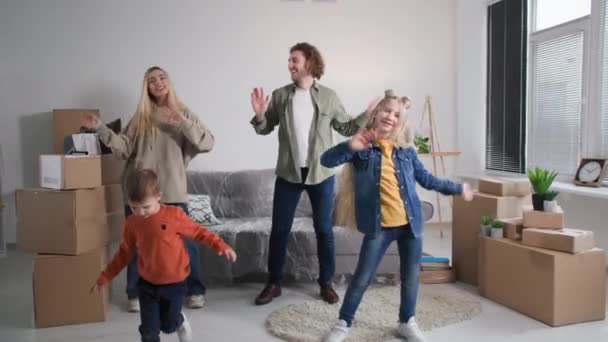 息子と娘を持つ幸せな両親は引越しの日に一緒に踊るのが楽しいです 家やアパートの購入を祝う段ボール箱に囲まれた若い家族 — ストック動画
