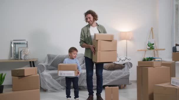 引っ越し後にポリエチレンで覆われた家具付きの新しいアパートに若いハンサムな父親と息子が段ボール箱で立って 残りの間に親と男の子の肖像画 — ストック動画