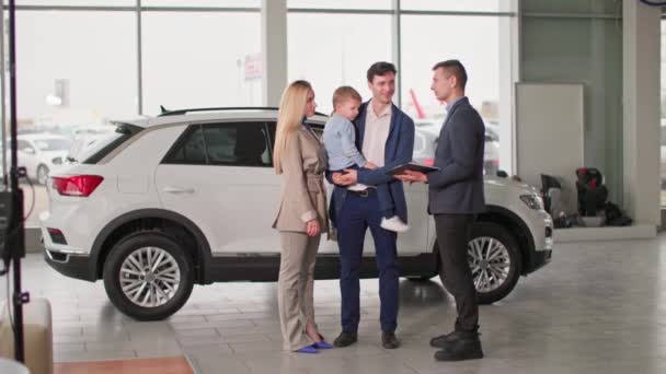 車の販売 幸せな夫と腕の中で息子と妻は 自動車センターで新しい自動車を選ぶことについてマネージャーと相談します — ストック動画