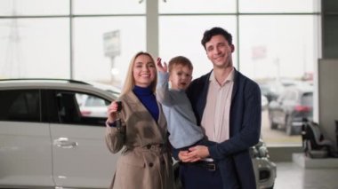 Otomobil fuarındaki aile, mutlu eşler, ellerinde yeni bir otomobilin anahtarları dururken, otomobil merkezinde durup gülümseyen ve kameraya bakan çocuklar.