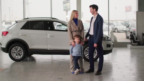 男性の子供を持つ若い家族の肖像は車のディーラーで新しい車を購入することを喜びます カメラでキーと笑顔を表示します — ストック動画