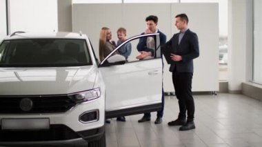 Araba alıyor, gülümseyen bir aile ve kollarında bir erkek çocuk bir otomobil galerisinde satıcıyla konuşuyor.