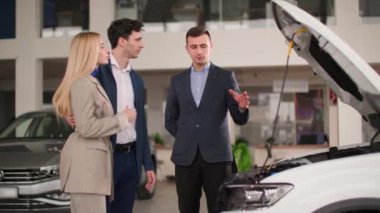 Genç eşler modern bir araç seçiyor, bir araba galerisinin profesyonel müdürü erkek ve kadın alıcılara alışveriş merkezindeki yeni bir otomobil modelinden bahsediyor