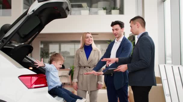 トランクの息子を持つ幸せな若い両親は自動車センターのセールスマネージャーと一緒に家族のための新しい車を選ぶ — ストック動画