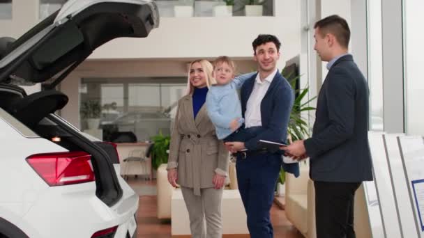 満足した男性と女性のバイヤー小さな男の子は家族の車を選択し マネージャは新しい自動車モデルを示し 自動車センターでトランクを開きます — ストック動画