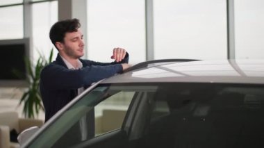 Genç ve başarılı bir adamın portresi otomobil galerisinden araba alırken seviniyor, parmağını kaldırıyor, gülümsüyor ve kameraya bakıyor.