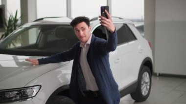 Yeni araba alan neşeli adam otomobil galerisinden yeni araba almaktan mutlu. Cep telefonuyla video çekiyor ve mutlulukla arabanın kaputunda yatıyor.