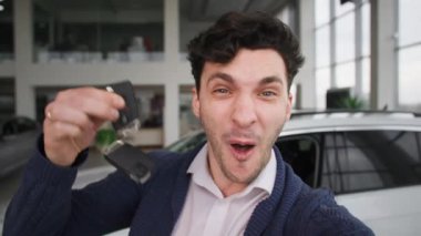 Ellerinde anahtarları olan genç ve mutlu bir adam, bir araba galerisinden yeni bir otomobil satın alırken telefonda bir video kaydediyor, kameraya bakıyor.