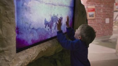 modern museum, little boy plays on a touch monitor and cleans a virtual rock from ground with his hands indoors
