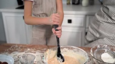 buns, female child together with caring mother prepare dough and mix wheat flour with eggs and milk in bowl with spoon on kitchen table, close-up