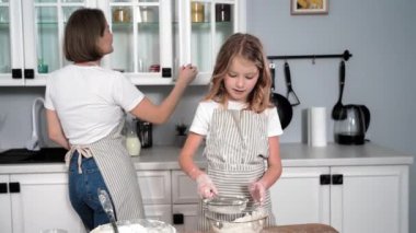 cooking with daughter, adorable female child preparing pastry dough and clapping hands with flour on background of mom with milk in hands