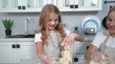 portrait of an adorable little girl with flour on face preparing dough together with mother in kitchen, smiling and looking at camera