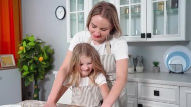 daughter-mother relationship, joyful girl together with woman in an apron roll out dough for cookies from wheat flour at table backdrop of kitchen
