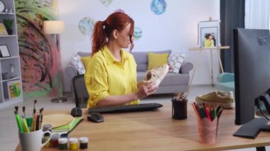 Ayakkabı tasarımı, elinde spor ayakkabıları olan genç yaratıcı kadın modern bir dairede otururken ayakkabı tasarımını yenilemenin yollarını arıyor.