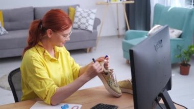 Ayakkabı tasarımları, gözlüklü kadın modern odada otururken renkli keçeli kalemlerle spor ayakkabılara moda baskısı çiziyor.