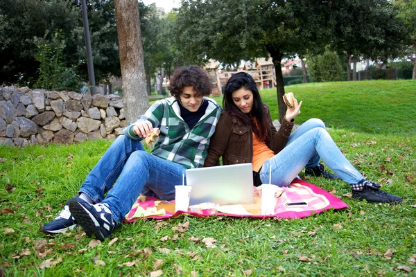 Happy couple watching pc while eating hamburger in park — Stock Photo, Image