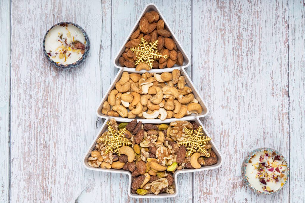 Mixed nuts and gold Christmas ornaments in a pine shape plate on light blue wooden background.
