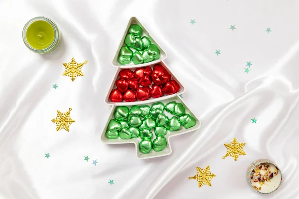 圣诞树形盘子里红绿相间的巧克力糖果 背景是白色缎子布 上面点着芬芳的蜡烛 — 图库照片