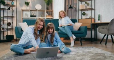 Aile eğlencesi konsepti. Sevimli, güler yüzlü, dost canlısı iki kız kardeşin yerde oturup dizüstü bilgisayar kullanması ve annelerine merhaba demek için el sallaması. Kanepede hangi kitap okunur?