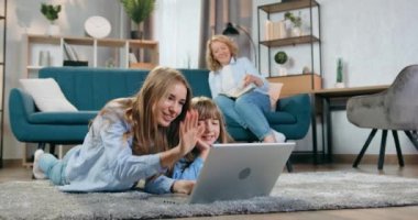 Sevimli, pozitif, mutlu, sarışın anne kanepede kitap okuyor ve yerde yatıp dizüstü bilgisayar kullanarak gülümseyen samimi büyük ve küçük kızlarına bakıyor.