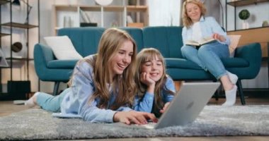 Güzel gülümseyen iki kız kardeş, en küçük ve en büyük iki kız kardeş, yerde yatar ve bilgisayar kullanırlar. Bu arada onların kaygısız anneleri oturma odalarında kanepede kitap okurlar.