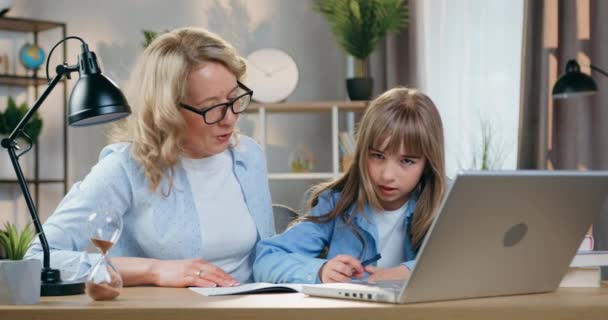 Miła pozytywna pewna siebie opiekuńcza blond matka w okularach pomaga swojej inteligentnej córce w odrabianiu lekcji, siedzi w miejscu pracy i czyta zadania na ekranie laptopa, zbliżenie — Wideo stockowe