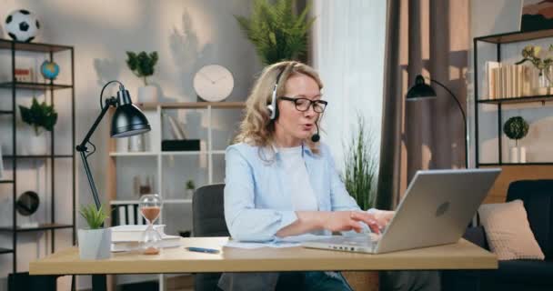 Attractive confident skilled modern blond woman in headset talking in mic simultaneously typing on laptop,working distantly from home office — Stock Video