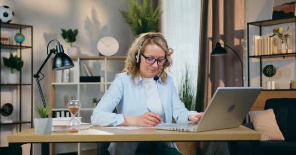 Hem arbete koncept där vacker säker ansvarsfull kompetent kvinna i headset arbetar med dokument och information på laptop på arbetsplatsen i mysiga rum — Stockvideo