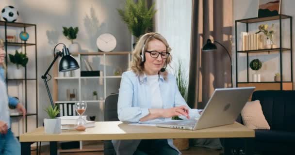 Good-looking confident positive skilled woman in headset working at home on computer when her playful active daughter running up to her and give huggs