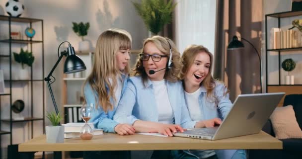 Schöne selbstbewusste erfolgreiche Frau mit Kopfhörern, die zu Hause am Computer arbeitet, wenn ihre jüngeren und ältesten Töchter auf sie zulaufen und reden — Stockvideo