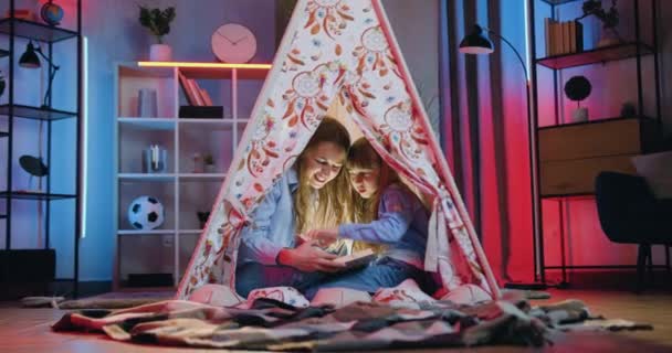 Aantrekkelijk glimlachen gelukkig vriendelijk twee verschillende leeftijden zussen zitten in wigwam 's nachts en het lezen van interessante boek — Stockvideo
