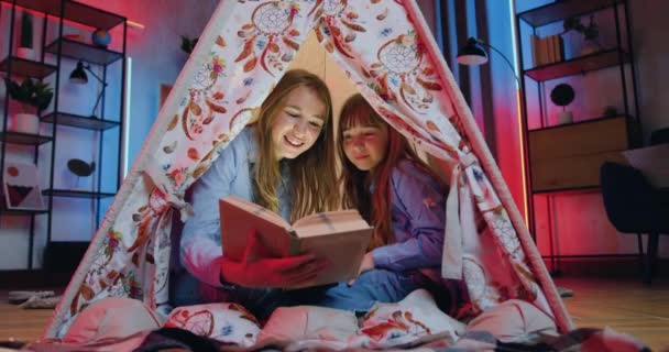 Close-up de simpática alegre simpática duas irmãs de diferentes idades que gastam seu lazer conjunto em casa e ler livro em barraca decorativa incrível à noite — Vídeo de Stock