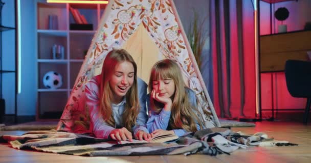 Liebenswert lächelnd glücklich freundlich zwei Schwestern unterschiedlichen Alters, die es sich auf Bettdecken im dekorativen Zelt gemütlich machen und über das Buch sprechen, das sie abends lesen — Stockvideo
