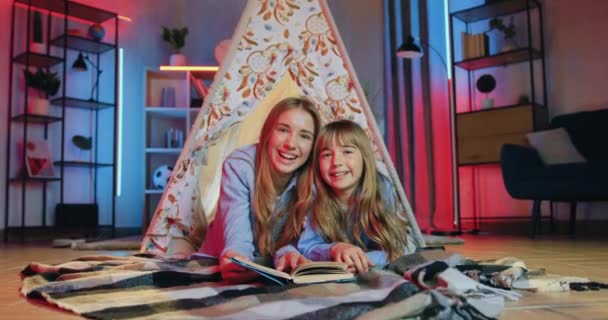 Nahaufnahme von zwei blonden Mädchen unterschiedlichen Alters, die glücklich lächelnd in die Kamera schauen, während sie abends im dekorativen Zelt zu Hause spannende Bücher lesen — Stockvideo