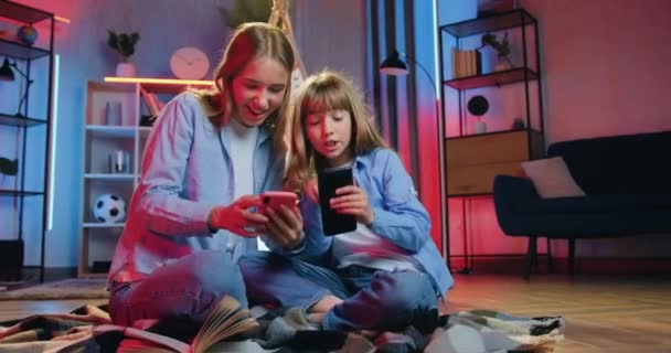 Camera shooting on beautiful cheerful emotional friendly two girls which having fun together at home in the evening and applying their phones — Stock Video