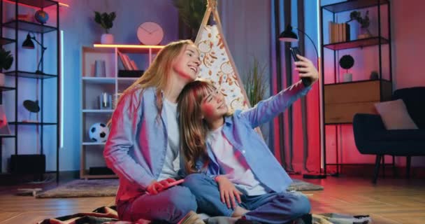 Vrije tijd concept waar mooie vrolijke vriendelijke verschillende leeftijden zussen het maken van selfie op de telefoon met grappige grimaces op nacht kamer interieur achtergrond — Stockvideo