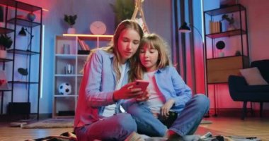 İyi görünümlü güler yüzlü mutlu iki farklı kardeş akşamları evdeki yorganın üzerinde oturuyor ve telefon kullanarak ortak fotoğrafları, ön görüntüyü izliyorlar.