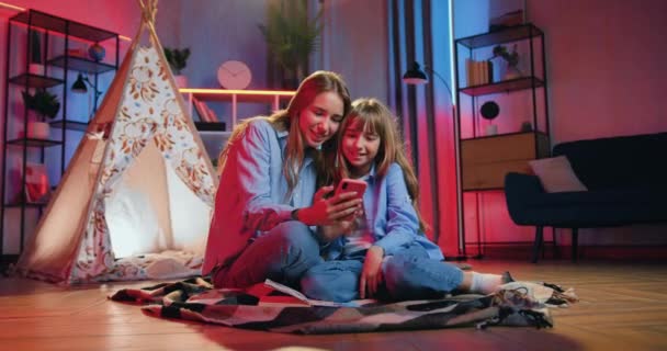 Familie idylle waar leuk glimlachen gelukkig zorgeloos blond twee inheemse verschillende leeftijden zussen samen plezier hebben in de avond thuis op dekens op de vloer met smartphone — Stockvideo