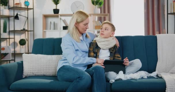 Attractive positive caring blond mother spending time together with her sick son with flu at home and they playing games on tablet pc,healthcare and family leisure concept — Stock Video