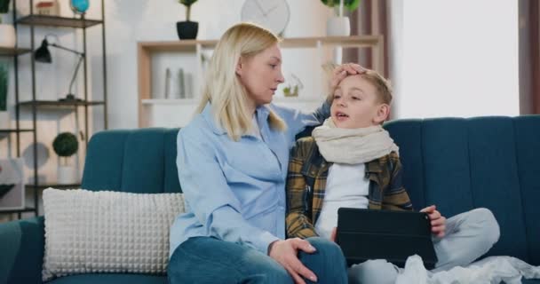 Good-looking caring loving woman sitting near her sick positive son on the couch and calming him,stroking on head,they have joint conversation ,front view — Stock Video