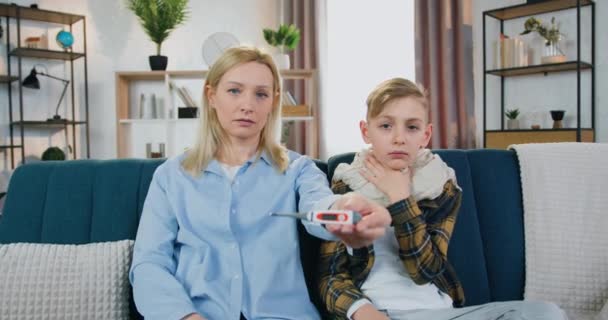 Porträt der attraktiven aufgebrachten blonden Frau, die die Temperatur auf dem Thermometer ihres kranken müden Teenie-Sohnes zeigt, der neben ihr auf dem heimischen Sofa sitzt, Gesundheits- und Kinderbetreuungskonzept — Stockvideo