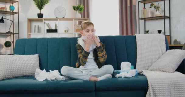 Хвороба та концепція домашнього лікування, де сумний хлопчик-підліток з грипом дме носом за допомогою серветки та кашлю, сидить на дивані, вид спереду, повільний рух — стокове відео